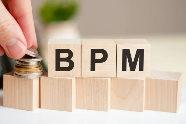 The word BPM written on wood cubes. A man's hand places the coins on the surface of the cube. Green plant in a flower pot on the background. Business and finance concept. Business Process Management