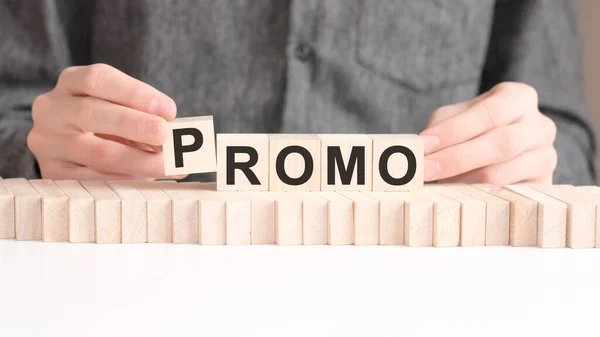 The hand puts a wooden cube with the letter P from the word Promo. The word is written on wooden cubes standing on the white surface of the table.