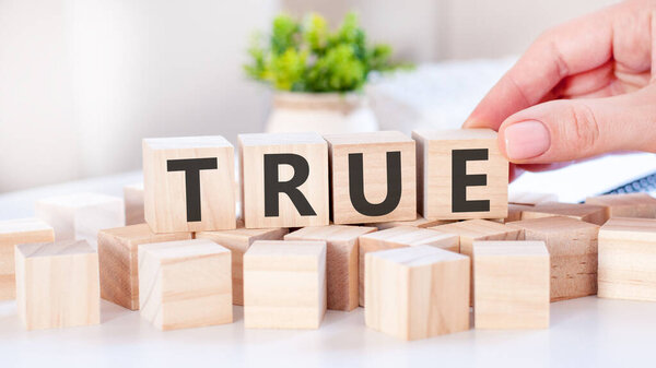 The hand puts a wooden cube with the letter E from the word True. The word is written on wooden cubes standing on the white surface of the table. Communication and business concepts