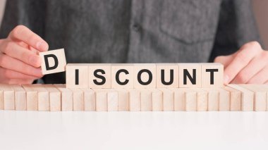 El, İndirim kelimesinin D harfinin olduğu tahta bir küp koyuyor. Kelime, masanın beyaz yüzeyinde duran tahta küplere yazılmıştır..