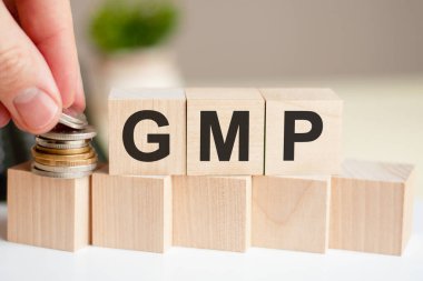 Tahta küplere GMP yazıyor. Bir adamın eli sikkeleri küpün yüzeyine yerleştirir. İş ve finans kavramı. GMP, iyi üretim uygulamasının kısaltılmışı