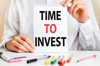 Beyaz gömlekli bir adam elinde bir kağıt parçası tutuyor: Yatırım yapma zamanı. Masada çok renkli kalemler ve tablet var. İş ve eğitim kavramı.