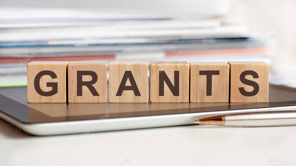 The word grants is written on wooden cubes standing on a notepad, in the background a stack of documents, selective focus. Can be used for business, education, financial, medical concept.