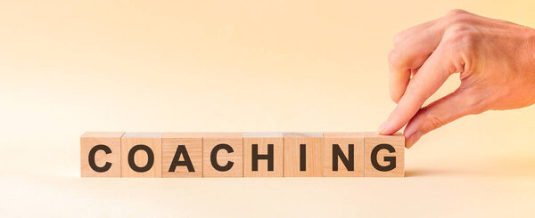 Рука кладет деревянный куб с буквой G от слова Коучинг. Слово написано на деревянных кубиках, стоящих на желтой поверхности стола. Может быть использована для концепции бизнеса и образования.