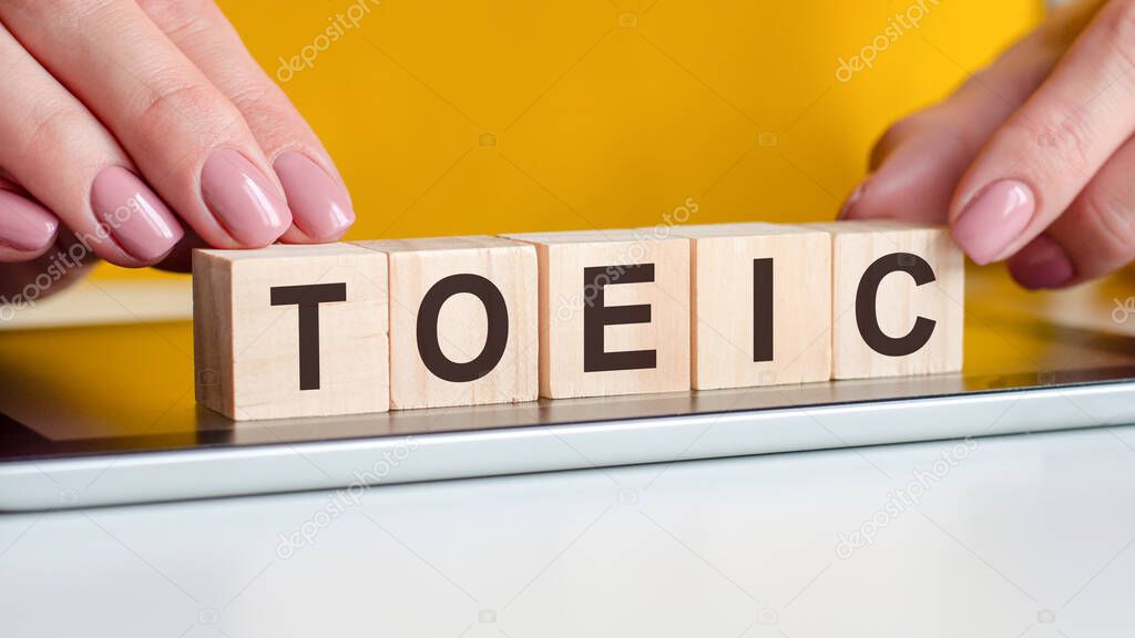 Women hands puts a wooden blocks with the letters toeic on the surface of the notepad. Can be used for education concept. Yellow background. TOEIC - test of english for international communication