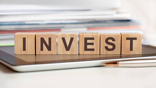 Слово INVEST написано на деревянных кубиках, стоящих на планшете, на заднем плане стопка документов, избирательный фокус. Может использоваться для бизнеса, образования, финансов, маркетинга.