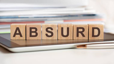 The word absurd is written on wooden cubes standing on a tablet, in the background a stack of documents, selective focus. Can be used for business, education, financial, marketing concept.
