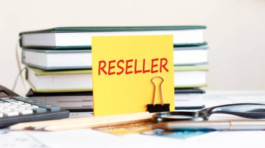 Yellow piece of paper with text reseller stands on a clip for papers on the desk against the background of books stacked, calculator, credit cards. Business and financial concept. Selective focus.