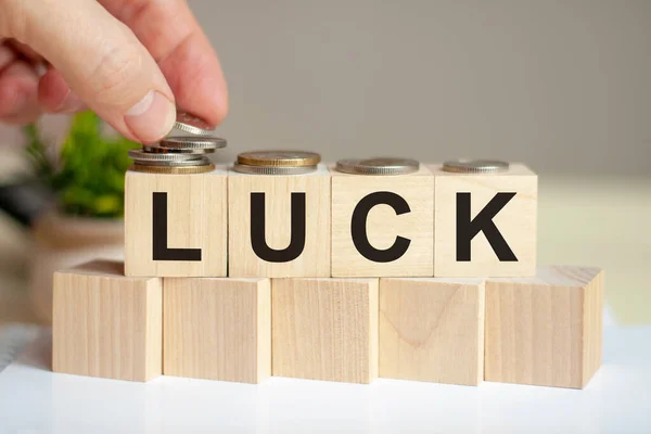 The word luck written on wood cubes. A man\'s hand places the coins on the surface of the cube. Green potted plant on the background. Business and finance concept.