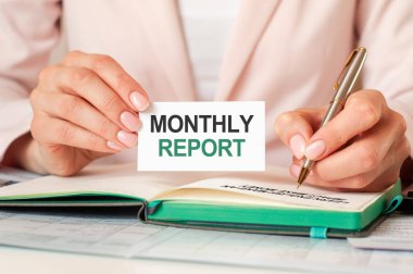woman writes in a notebook with a silver pen and hand hold card with text: monthly report. pink background, front view. business and education concept.