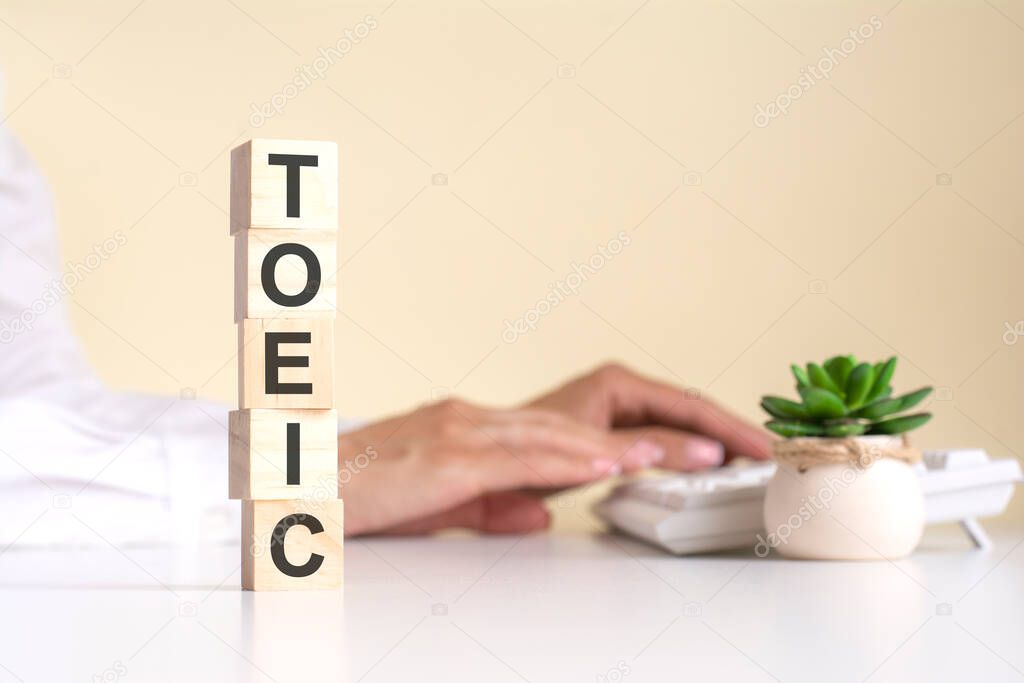 pyramid of wooden cubes with the word TOEIC in the foreground, in the background - business woman works on the keyboard in a light office, free space. selective focus