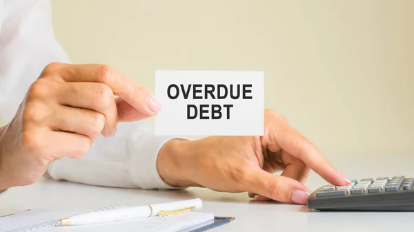overdue debt, message on business card shown by woman pressing calculator key at workplace in light office, selective focus, business and financial concept