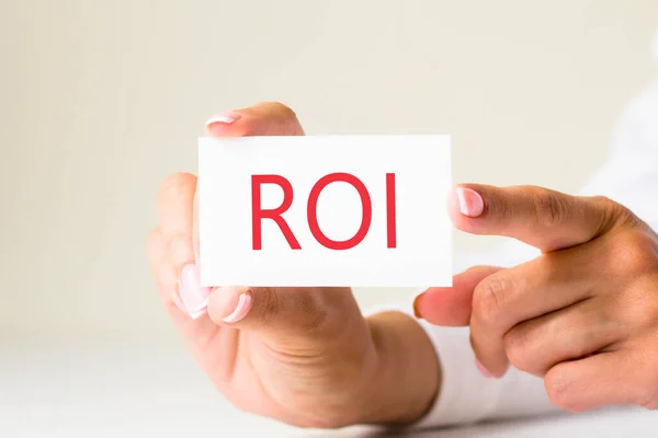 female hands hold card paper with text ROI - RETURN ON INVESTMENT - on a white light background. business and finance concept