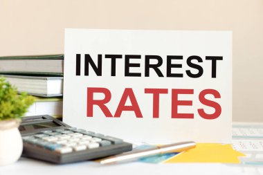 Metin faiz oranları olan beyaz kart, dizilmiş kitapların, hesap makinesinin, kalemin, yeşil saksı bitkisinin arka planında duruyor. İş ve finansal konsept. Seçici odak.