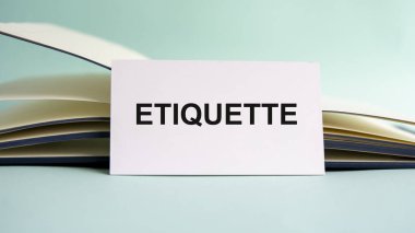 Üzerinde ETIQUETTE metni olan beyaz bir kartvizit açık bir günlüğün arka planında bir masanın üzerinde duruyor. Odaklanmamış.