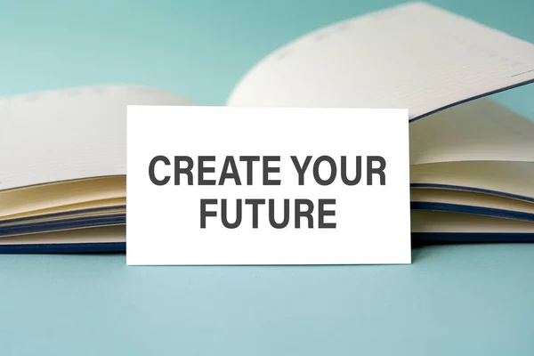 Um cartão branco com CREATE YOUR FUTURE texto está em uma mesa contra o fundo de um diário aberto. Desfocado — Fotografia de Stock