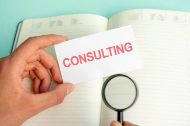 Bir adamın elinde beyaz bir kart var. Üzerinde açık bir kağıt defter ve siyah çerçeveli bir büyüteç var.