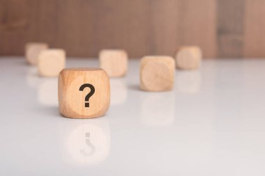 Üzerinde soru işareti olan tek bir tahta blok, arka planda bulanık tahta küplerle yansıtıcı beyaz bir masanın üzerinde duruyor. Gizemi, soruşturmayı, bilinmeyeni taşır.