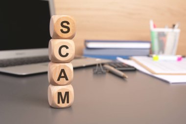 SCAM sözcüğünü oluşturmak için dizüstü bilgisayarı, belgeleri ve ofis malzemelerini arka planda bulanıklaştıran dört tahta blok yığılmış. Görüntü, finansal dolandırıcılık, aldatma, risk ve ihtiyatı çağrıştırıyor..