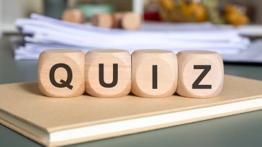 Üzerinde Q-U-I-Z harfleri olan dört doğal tahta küp bulanık dökümanların önünde kapalı bir deftere yerleştirilmiş. Sahne bir ofiste ya da akademik ortamda sınav, sınav, bilgi kontrolü, test ya da anket yapılmasını önerir