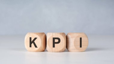 Arka arkaya dizilmiş üç tahta küp ve hafif bir masa yüzeyindeki K-P-I harfleri yumuşak nötr bir arkaplana karşı. Temiz, asgari bileşim temel performans göstergelerini, ölçümleri, iş hedeflerini, analizleri, başarı ölçümlerini ve şirketleri iletir