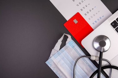 Tıbbi steteskop, dizüstü bilgisayar klavye, kırmızı kredi kartı, takvim ve koyu gri arka planda cerrahi maske ile çalışma alanının en üst görüntüsü. Sağlık sigortası ödemesi kavramı, çevrimiçi doktor randevu defteri, teletıp giderleri ve sağlık hizmetleri