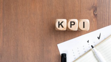 KPI Anahtarı Performans Göstergesi 'ni koyu ahşap arka planda heceleyen tahta küplerin üst görünümü. Kompozisyonda beyaz bir işaret ve işaretli bir takvim yer alıyor. İş analizleri, kurumsal habercilik, verimlilik takibi için ideal
