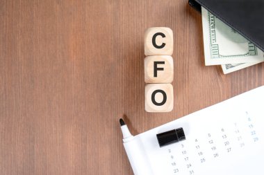 Koyu ahşap arka planda, dikey tahta bloklar halinde CFO (Mali İşler Müdürü) yazılıyor. Kompozisyonda içinde dolar banknotları olan bir cüzdan, işaretli bir takvim ve beyaz bir işaret var. Kurumsal yönetim kavramı, mali rapor