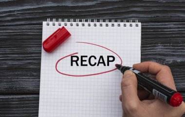 Bir kadının eli beyaz bir çarşafa siyah ahşap arka planda RECAP kelimesini yazar.