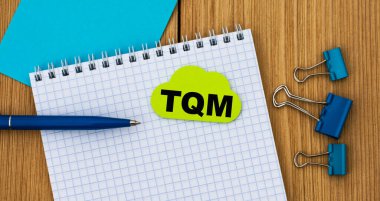 TQM (Toplam Kalite Yönetimi) - tahta bir arka planda bir deftere kalem ve kıskaçlarla yazılmış bir kelime. İş ve finans kavramı