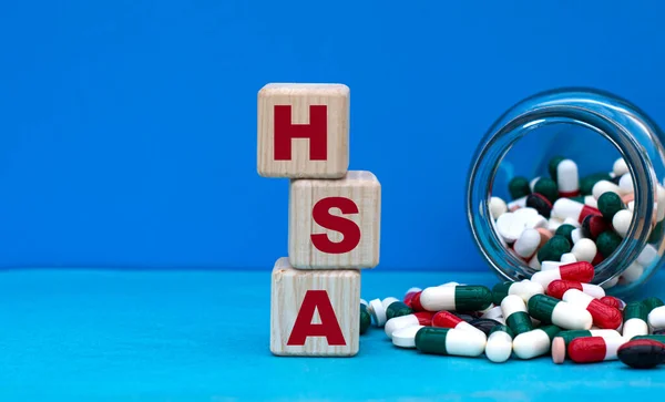 HSA (Health Savings Account) word on cubes on a blue background with a jar of tablets. Medical concept.