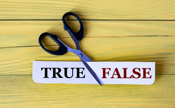 Scissors on a wooden yellow background cut paper with the words TRUE and FALSE. Info concept