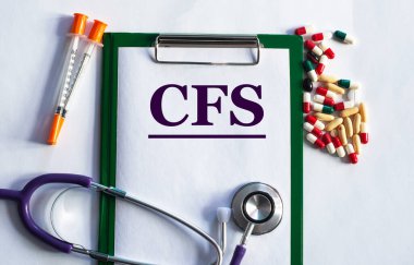 Not defterinde steteskop ve haplarla dolu bir masada CFS (Kronik Yorgunluk Sendromu) kelimesi vardı.