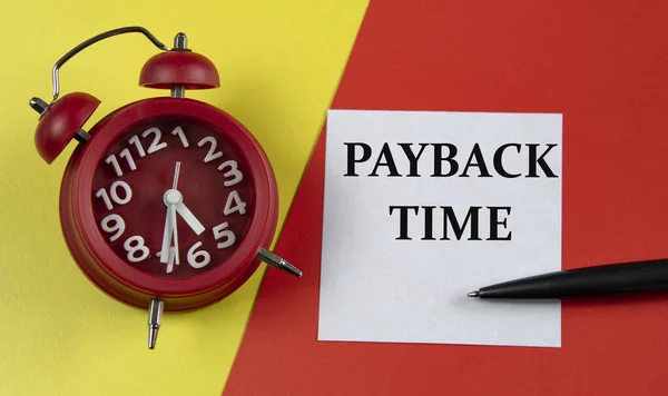 PAYBACK TIME - words on a white sheet on a multi-colored background with an alarm clock and a pen. Business concept