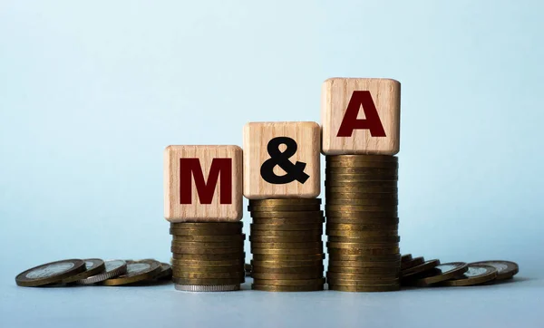M and A (Mergers and Acquisitions) - acronym on wooden cubes. Which stand on stacks of coins on a light background. Business concept