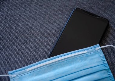 Yakın plan koruyucu tıbbi maske, gri desenli kumaşta siyah telefon. Fotokopi alanı olan duvar kağıdı ve arkaplan. İç kısım için kalın gri malzeme. Coronavirus konsepti. Yeni gerçeklik.