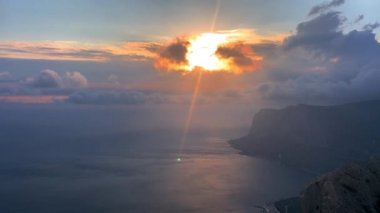 Denizin üzerinde gün batımı. Deniz kıyısında hızlı akan bulutlar. Güçlü rüzgar su yüzeyinde bulutlar savurur. Deniz güneşi yansıtır. Gerçek zamanlı video. Doğal arka plan. Güneş, deniz ve dağ manzarası.