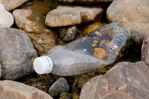 A transparent plastic bottle with a white cap lies on the river bank among the stones.