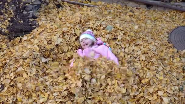 Het meisje ligt in een stapel gevallen bladeren en maakt een engel. Herfst op de Russische werf — Stockvideo