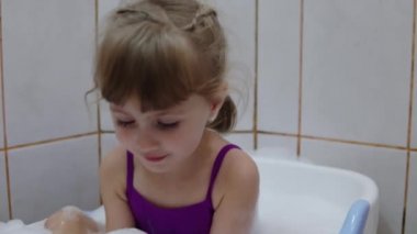 Bir kız bebek banyosunda banyo yapar ve ellerindeki köpüğü temizler.