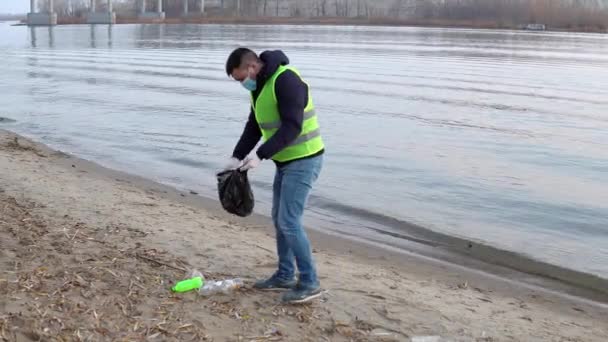 Een vrijwilliger met een masker en handschoenen loopt langs het strand en verzamelt afval in een tas. Milieuverontreiniging — Stockvideo