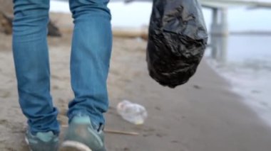 Bir gönüllü kumsalda yürüyor ve çöpleri yakın plan çekimde topluyor. Çevre kirliliği