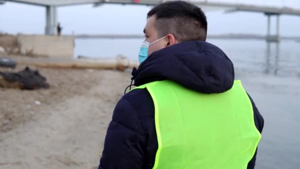 Um voluntário usando uma máscara e luvas caminha pela praia e coleta lixo em um saco. Poluição ambiental — Vídeo de Stock