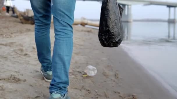 Um voluntário caminha ao longo da praia e coleta lixo em um saco de close-up. Poluição ambiental — Vídeo de Stock