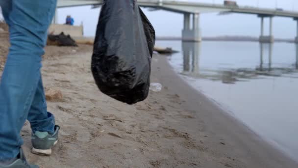 Um voluntário caminha ao longo da praia e coleta lixo em um saco de close-up. Poluição ambiental — Vídeo de Stock