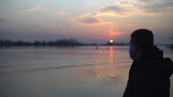 Een man met een masker staat bij de rivier, het meer en kijkt naar de zonsondergang — Stockvideo
