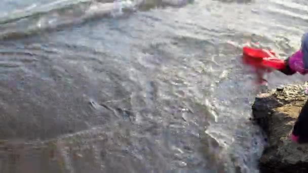 子供は川のそばの石の上に立っておもちゃのシャベルで遊ぶ — ストック動画