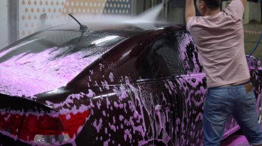 Adam arabayı yıkar. Yüksek basınçlı suyla pembe köpüğü temizler. 4k