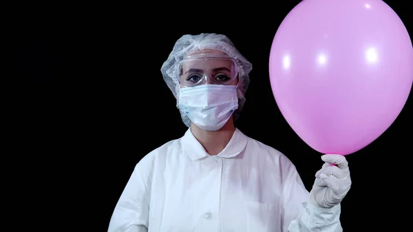 A nurse in protective gear holds a pink balloon in her hand and pierces it with a syringe 4k