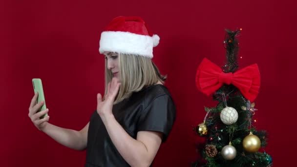 Kobieta w kapeluszu Świętego Mikołaja na czerwonym tle mówi przez wideo połączenie w pobliżu choinki — Wideo stockowe